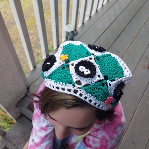 May include: A handmade crochet hat with a patchwork design in green, white, and black squares. The hat is decorated with small, colourful floral accents. A unique accessory.