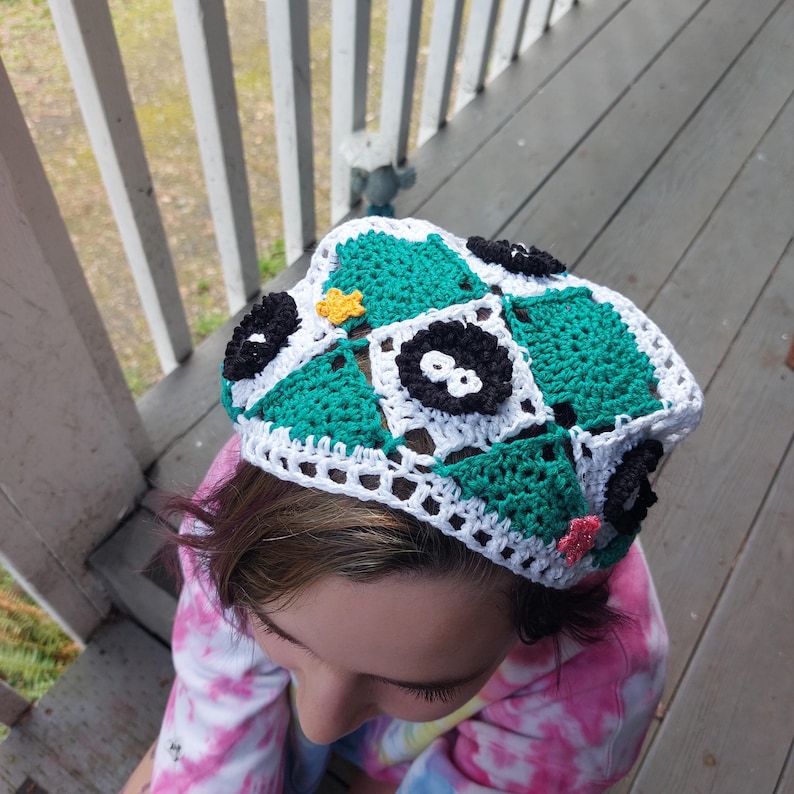 May include: A handmade crochet hat with a patchwork design in green, white, and black squares. The hat is decorated with small, colourful floral accents. A unique accessory.