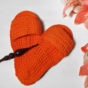 May include: A pair of orange crocheted slippers in the shape of a heart. A brown crochet hook is lying on top of the slippers.