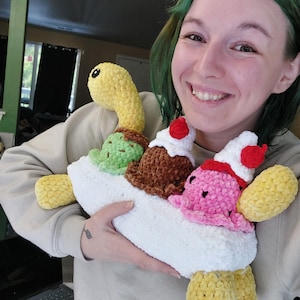 May include: A handmade, plush ice cream sundae with a yellow turtle head and feet. The sundae features scoops of green, brown, and pink ice cream, topped with red cherries and whipped cream, all nestled in a white crocheted boat.