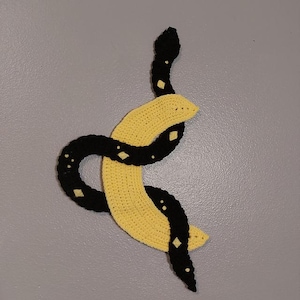 Puede incluir: Arte de pared tejido a ganchillo que representa una serpiente negra enrollada alrededor de una luna creciente amarilla. La serpiente tiene detalles en forma de diamante y la luna tiene una superficie texturizada. La obra de arte se muestra sobre un fondo gris.