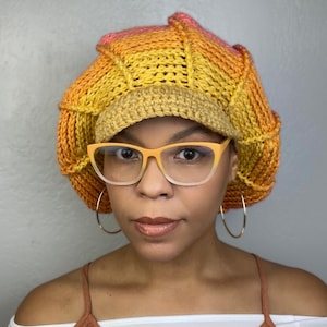 May include: A crocheted hat with a yellow, orange and pink ombre colour scheme. The hat has a wide brim and is worn by a person with glasses.