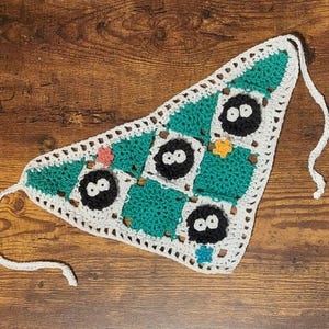 May include: Handmade crochet bandana featuring a patchwork design with teal and white squares. Each square has a black character with white eyes. The bandana has a white border and ties.