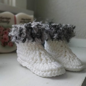 May include: A pair of handmade white crochet booties with a fluffy gray and black trim around the ankles. The booties are made with a thick yarn and have a textured appearance, perfect for keeping feet warm.