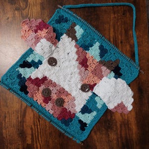 May include: A handmade crochet cow wall hanging with a turquoise border and a matching strap. The cow's face is white with brown button eyes and colorful patches of pink, peach, and brown. The piece is designed to be hung on a wall.