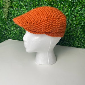 Op de afbeelding: Een oranje gebreide newsboy-pet met een platte bovenkant en een rand.
