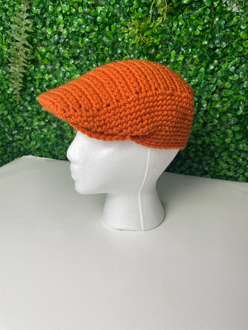 Op de afbeelding: Een oranje gebreide newsboy-pet met een platte bovenkant en een rand.