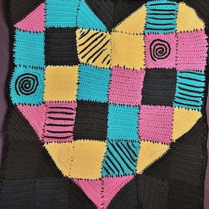May include: A handmade crochet pillow with a heart design. The heart is composed of various colored squares: black, teal, pink, and yellow. Some squares have black spiral or striped accents. The pillow is framed by a black border.