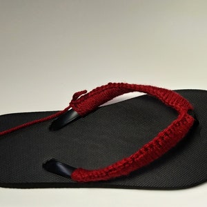 May include: A black flip-flop with a red crocheted strap. The strap is attached to the flip-flop with a black string.