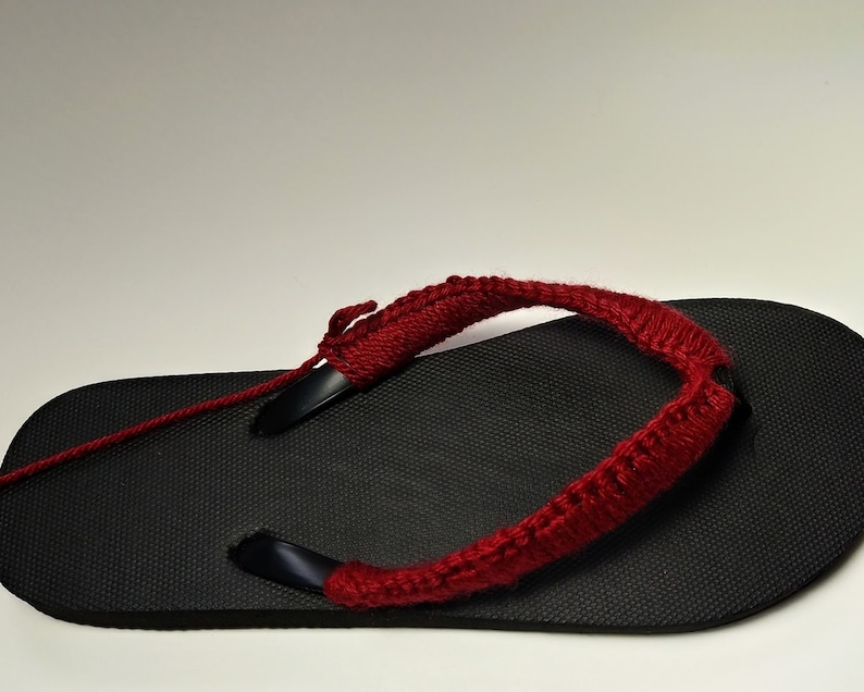 May include: A black flip-flop with a red crocheted strap. The strap is attached to the flip-flop with a black string.