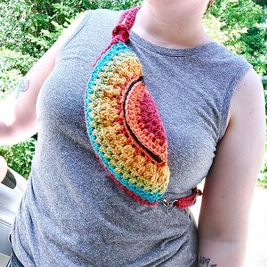 Op de afbeelding: Handgemaakte gehaakte heuptas in een regenboog van kleuren, waaronder rood, oranje, geel, groen en blauw. De tas heeft een zwarte rand en een bijpassende riem. De riem is verstelbaar en heeft een metalen sluiting.