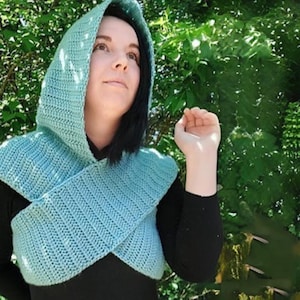 Puede incluir: Un conjunto de capucha y bufanda de ganchillo azul claro. La capucha está unida a una bufanda que cruza el pecho. El conjunto está hecho de un hilo suave y texturizado. La persona lleva una camiseta negra de manga larga.