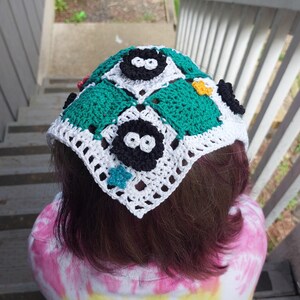 May include: A handmade crochet head covering with a patchwork design. The squares are white, green, and black, with cartoon characters and star accents. The head covering is worn over brown hair and a tie-dye shirt.