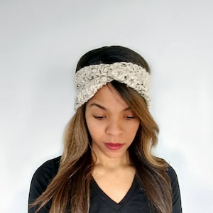 May include: A beige crocheted headband with a twisted knot design. The headband is worn by a person with long brown hair.