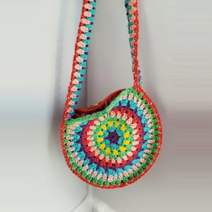 May include: A handmade crochet bag in a circular design, featuring a vibrant mix of colours. The bag includes red, green, blue, yellow, and purple. The strap and trim are red. The bag is suspended against a white wall.