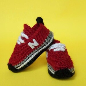 May include: A pair of red crocheted baby shoes with white laces and a black and white trim. The shoes have a white "N" on the front.