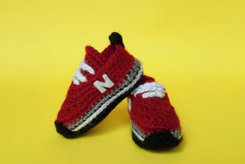 May include: A pair of red crocheted baby shoes with white laces and a black and white trim. The shoes have a white "N" on the front.