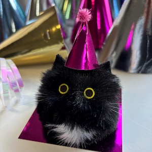 May include: A black cat ornament with yellow eyes and a white chest patch, wearing a magenta party hat with a pink pom-pom. Other metallic party hats in gold, silver, and pink are in the background.