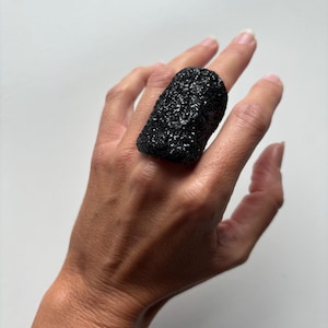 May include: A chunky black ring with a textured, glittery finish. The ring has a rectangular shape and is a prominent accessory. The ring's design suggests it is a statement piece, possibly made from a solid material.