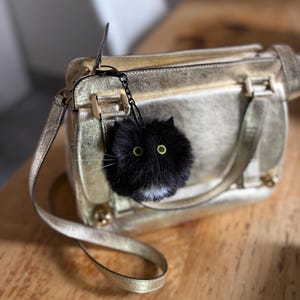 May include: A metallic gold handbag with a structured rectangular shape and a long shoulder strap. Attached to the bag is a round, fluffy black cat-shaped charm featuring two bright yellow eyes, pointed ears, and a small white patch on its chest.