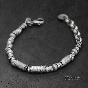 May include: A silver bracelet with a variety of textured beads and cylindrical elements. The bracelet features a lobster clasp closure and a mix of smooth and patterned surfaces. The jewelry is displayed on a dark background.