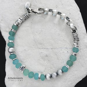 May include: A silver bracelet featuring seafoam green beads and textured silver accents. The bracelet includes a variety of bead shapes and sizes, with a secure clasp. The jewelry is displayed on a white, textured surface.