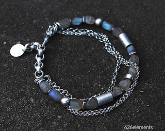 Puede incluir: Una pulsera de plata en capas con una mezcla de pequeñas piedras preciosas grises y azules irregulares y eslabones de cadena de plata. La pulsera tiene un pequeño dije redondo de plata y detalles cilíndricos de plata. Joyas.