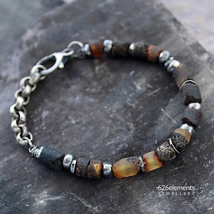 Puede incluir: Una pulsera artesanal con cuentas de color marrón oscuro y ámbar, intercaladas con pequeñas cuentas plateadas. La pulsera se asegura con una cadena y un cierre plateados. La joya se exhibe sobre una superficie gris texturizada.