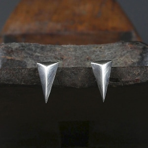 May include: A pair of silver-coloured stud earrings, each shaped as a three-dimensional triangle with a pointed tip. The metal surface features a brushed, textured finish with visible fine lines, creating a metallic sheen across the geometric form.