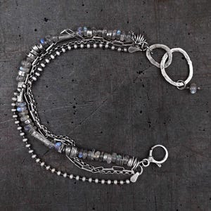 Könnte beinhalten: Ein mehrsträngiges Armband mit oxidierten silberfarbenen Metallketten, kleinen runden Metallperlen und facettierten grauen Labradoritsteinen mit blauem Schimmer. Das Schmuckstück enthält zwei gehämmerte ovale Glieder und einen Karabinerverschluss.