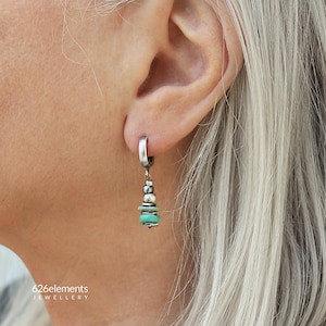 May include: A silver hoop earring featuring a dangling charm composed of small metallic beads and flat, circular turquoise-coloured stones. The earring is worn in an earlobe. The text 626elements JEWELLERY is visible in the bottom left corner.
