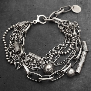 Könnte beinhalten: Ein mehrsträngiges silberfarbenes Armband mit einer Mischung aus strukturierten Kettengliedern, kleinen kugelförmigen Perlen und zylindrischen Metallakzenten. Das Schmuckstück verfügt über einen Karabinerverschluss und einen runden Anhänger mit oxidiertem Finish.