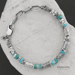 May include: A silver bracelet featuring a mix of textured cylindrical beads, small flat spacers, and vibrant turquoise stone chips. The piece includes a large oval link chain section and a lobster claw clasp with a small circular charm attached.