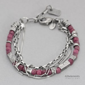 Op de afbeelding: Een armband met meerdere strengen met zilveren kettingen en donkerroze edelsteen kralen. De armband heeft verschillende kettingstijlen, waaronder een delicate kralenketting en een dikkere gourmetteketting. De sluiting en verbindende elementen zijn zilver.