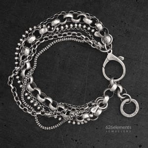 Puede incluir: Una pulsera de plata de varias hebras con diversos estilos de cadena, incluyendo eslabones rolo, de bola y texturizados. La pieza incluye un gran cierre de mosquetón y un dije circular con una textura fina y punteada. El texto 626elements JEWELLERY es visible.