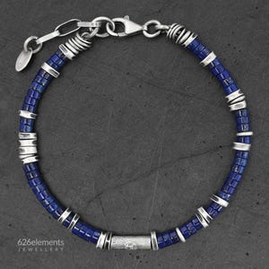Op de afbeelding: Een armband met kleine, cilindervormige blauwe lapis lazuli kralen afgewisseld met getextureerde zilverkleurige metalen tussenstukjes en een centrale zilverkleurige buiskraal. Het heeft een karabijnsluiting en een 626elements-label.