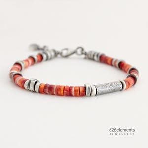 May include: A beaded bracelet featuring red and orange spiny oyster shell beads, interspersed with silver-colored metal accents. A decorative silver tube is centered on the bracelet. The bracelet is fastened with a silver clasp. The jewelry is from 626elements.