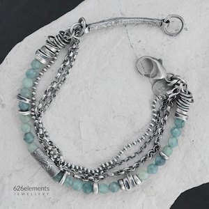 Puede incluir: Una pulsera plateada de múltiples hilos con cuentas azul claro y varios estilos de cadena. La pulsera presenta un cierre decorativo y una barra texturizada. La marca "626elements JEWELLERY" es visible.