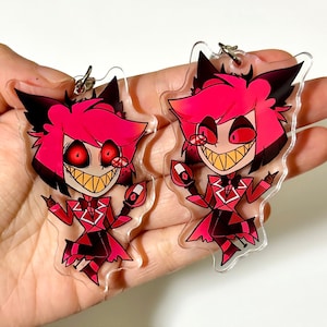May include: Two red and black cartoon character keychains with a devilish grin. The characters have red hair, black eyes, and are wearing a red suit with a white collar and bow tie.