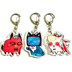 May include: Three colorful cartoon cat keychains with silver key rings. The first keychain features a red cat with horns and black eyes. The second keychain features a blue cat with a black TV screen for a head and a white bow tie. The third keychain features a white cat with a pink crown and red wings.