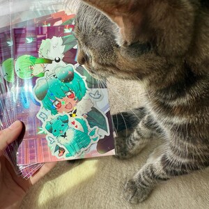 May include: A stack of colorful stickers featuring anime-style characters with vibrant teal and green hair, and a teal dog. The stickers are held by a hand, with a tabby cat in the foreground.