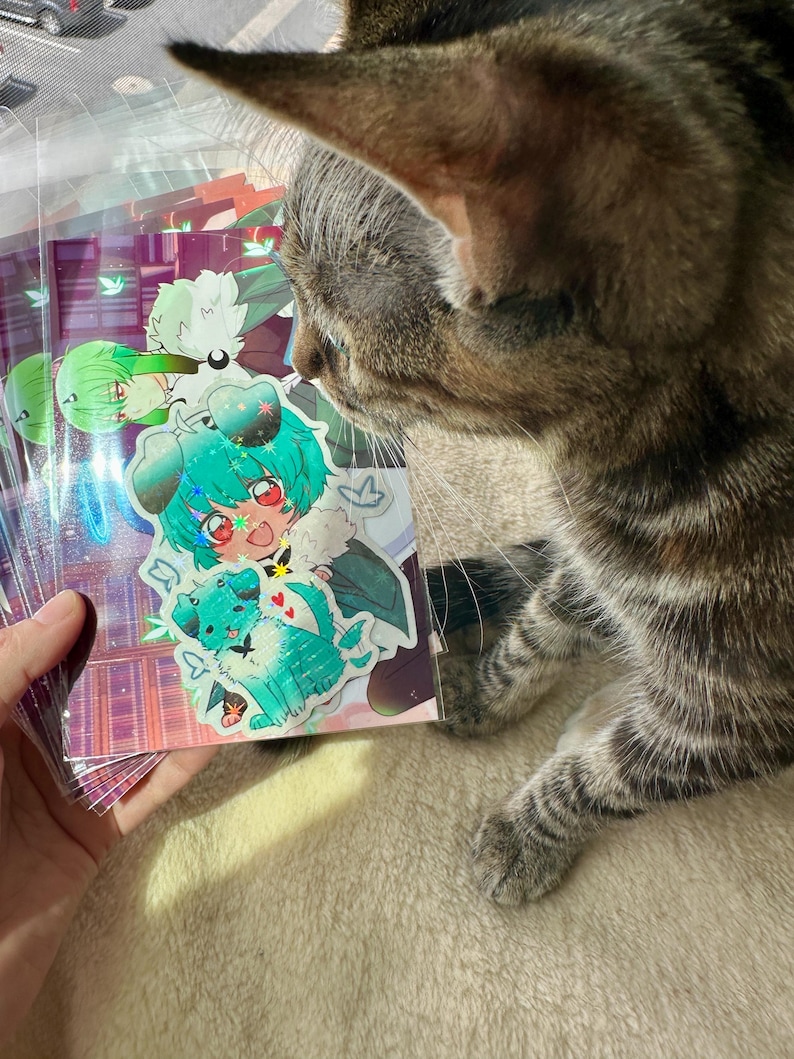 May include: A stack of colorful stickers featuring anime-style characters with vibrant teal and green hair, and a teal dog. The stickers are held by a hand, with a tabby cat in the foreground.