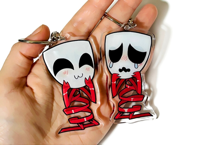 May include: Two clear acrylic keychain charms featuring cartoon faces. One charm has a happy face with red blush and the other charm has a sad face with red blush and tears. Both charms have red ribbon details.