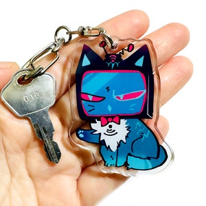 May include: A blue and black cat with a TV screen for a head, wearing a bow tie and a small antenna on top of its head. The cat is holding a paw up in the air. The cat is a keychain charm.