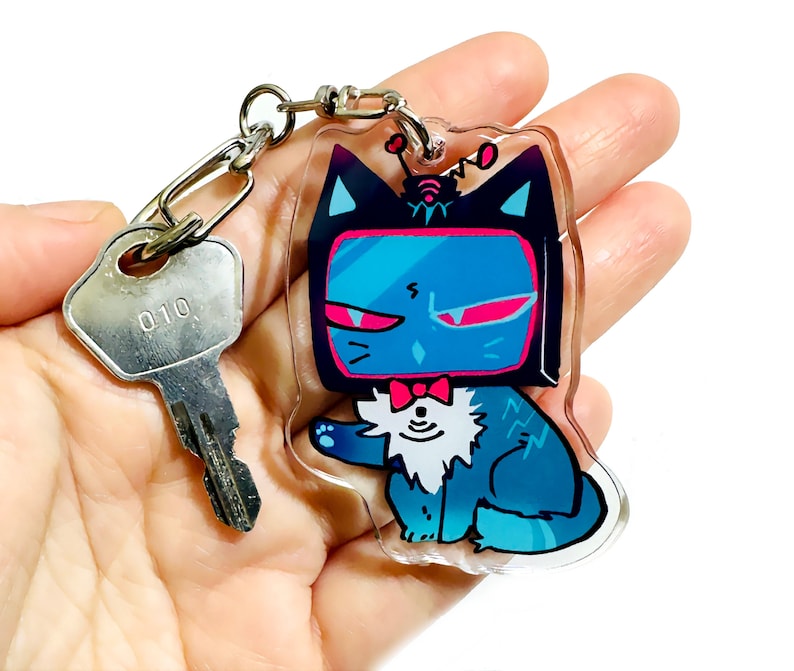 May include: A blue and black cat with a TV screen for a head, wearing a bow tie and a small antenna on top of its head. The cat is holding a paw up in the air. The cat is a keychain charm.