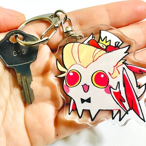 May include: A white and red cartoon character with a crown and a bow tie on a clear acrylic keychain. The keychain is attached to a silver key ring.
