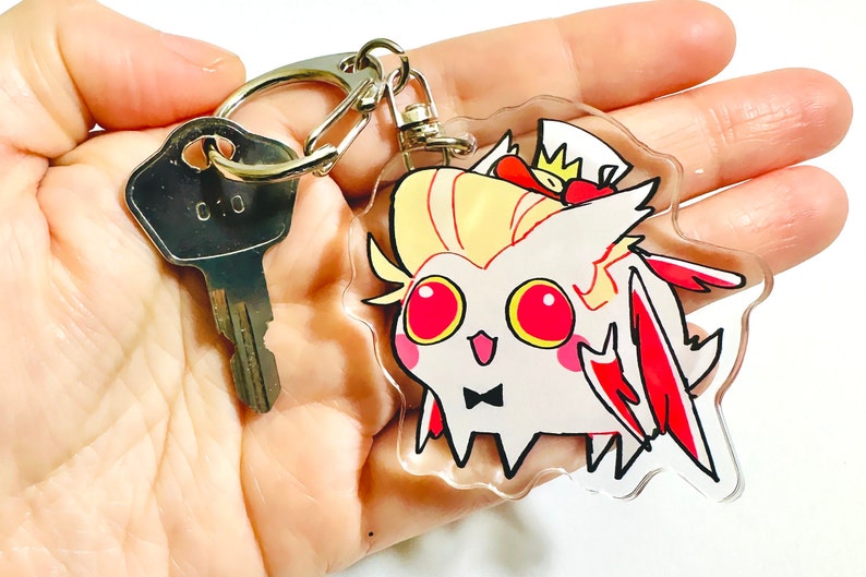 May include: A white and red cartoon character with a crown and a bow tie on a clear acrylic keychain. The keychain is attached to a silver key ring.