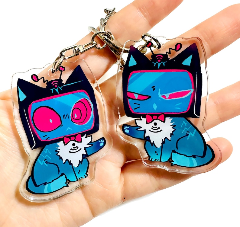May include: Two blue and black cat-shaped keychains with a television screen for a head. The television screens have pink and blue accents. The cats are wearing a bow tie and have a white fluffy collar.