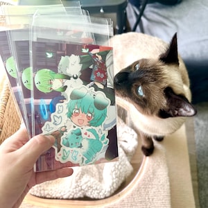 May include: A stack of clear plastic sleeves holding colorful anime-style art prints. The prints feature characters with green and blue hair, and a teal dog. A Siamese cat with blue eyes is in the background.