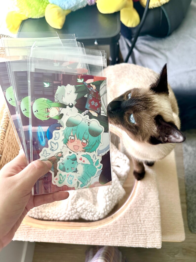 May include: A stack of clear plastic sleeves holding colorful anime-style art prints. The prints feature characters with green and blue hair, and a teal dog. A Siamese cat with blue eyes is in the background.